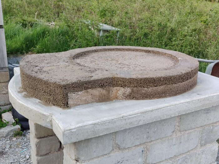 Rund betongplatta med grov yta, vilande på murad stenstruktur utomhus med vegetation i bakgrunden.