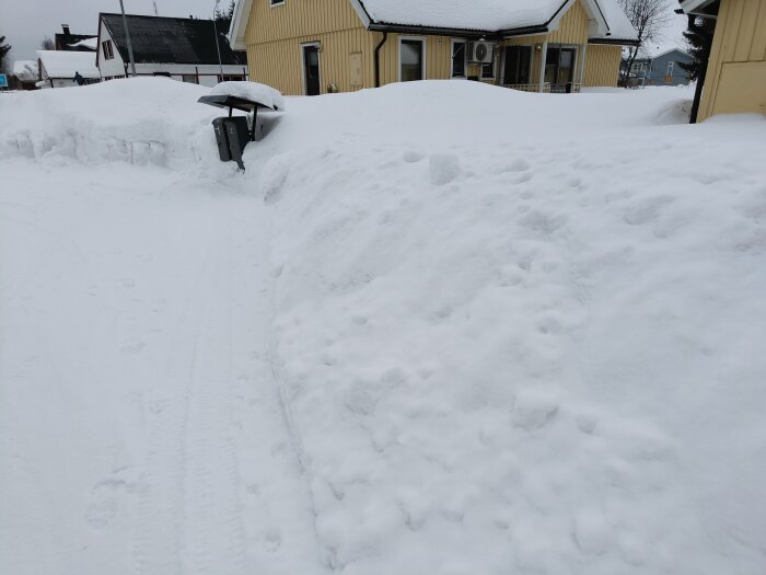 Snöig landsväg, högar av snö, gula hus, molnig himmel, synliga fotspår och däckspår.