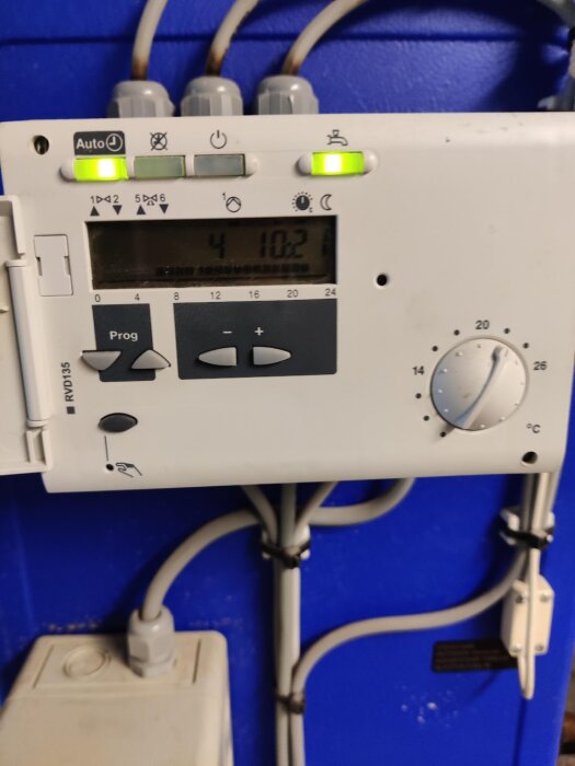 Programmerbar termostat för uppvärmningssystem, monterad på blå vägg. Kablar och rör anslutna, digital display visar tiden.