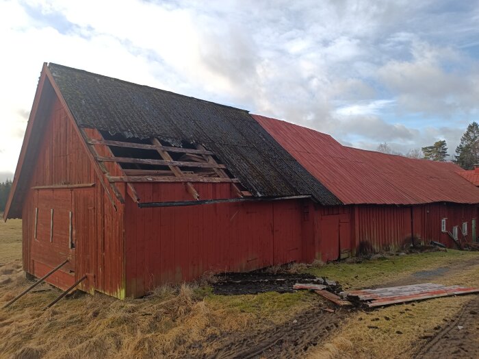 Stor röd ladugård med skadat tak, delar saknas, omgiven av gräs och träd.