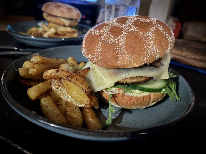 Hamburgare med ost och sallad, kryddiga potatisklyftor, mörkt tallrik, andra portion i bakgrunden, inomhus.