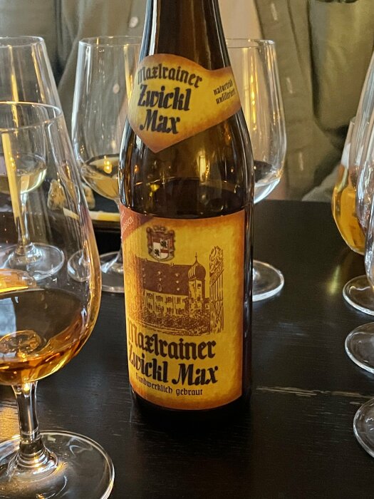 En ölflaska med etikett "Märzenbier Zwickl Max" framför tomma glas på ett mörkt bord.