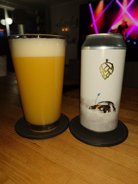 Ett glas öl och en ölburk på underlägg med inredning och suddig bakgrundsbelysning i rummet.