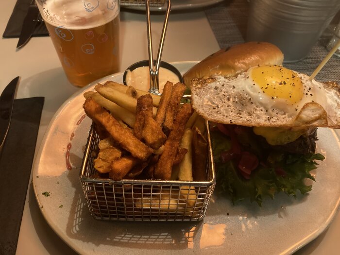 En hamburgare med stekt ägg, pommes frites i korg, ölglas och sås, restaurangmiljö.