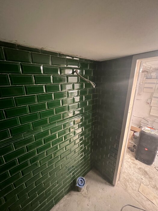 En halvfärdig grön kaklad vägg, kabel sticker ut, byggstök, säkerhetspanel, blå skyddsplast över golvbrunn.