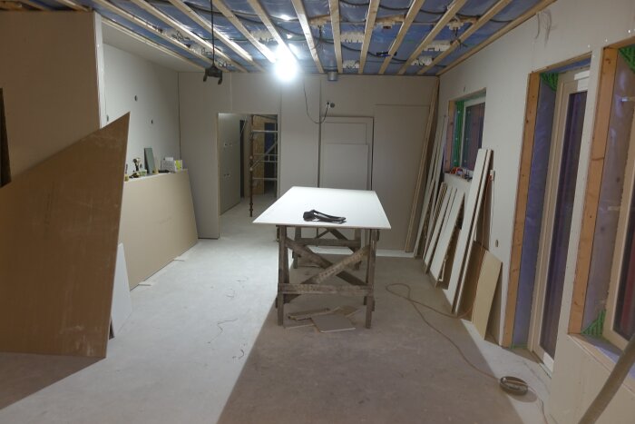 Under renovering, ouppklätt tak, isoleringsmaterial, byggmaterial, ensamt bord i mitten, arbetslampa, tomma väggar.