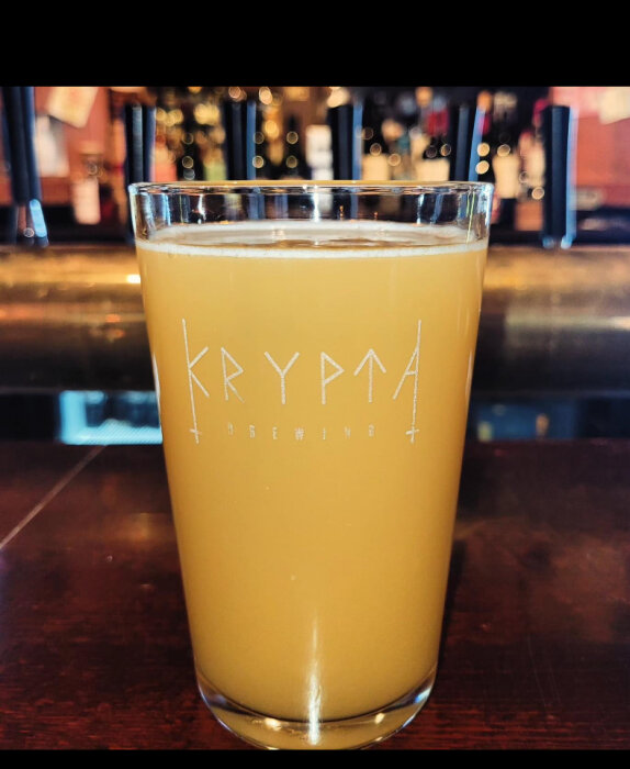Ett glas med grumlig, guldfärgad dryck på en bar, med suddig bakgrund och logotypen "Krypta Brewing".