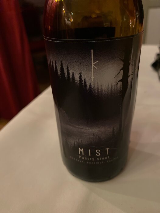 En flaska med etikett "MIST Pastry Stout" framför suddig bakgrund, mörk atmosfär med träd och dimma.
