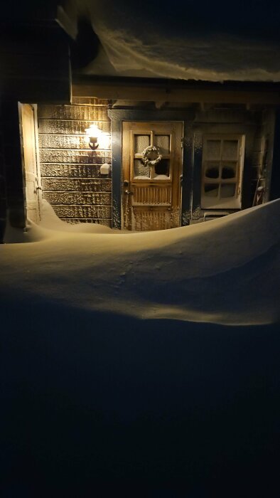 Husets entré täckt av tjockt snötäcke på natten, upplyst av vägglampa.