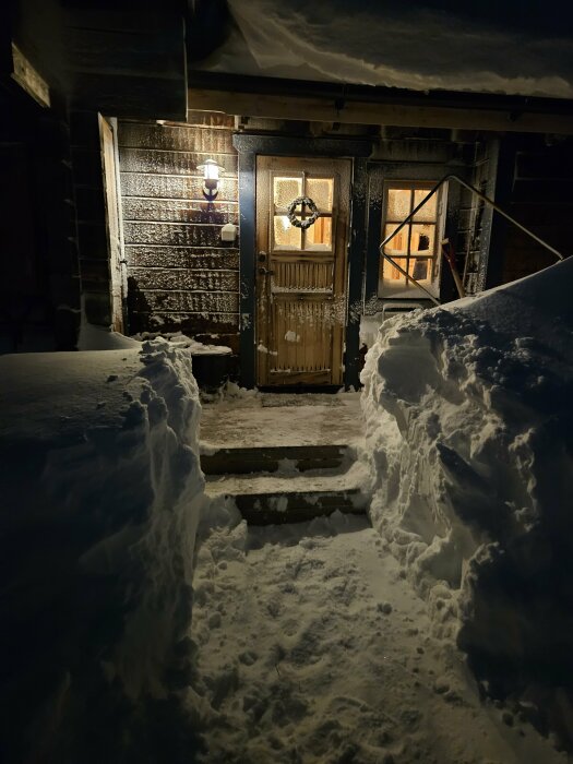 Snötäckt entré till hus på natten, upplyst av utomhuslampor, omgivet av höga snöväggar.