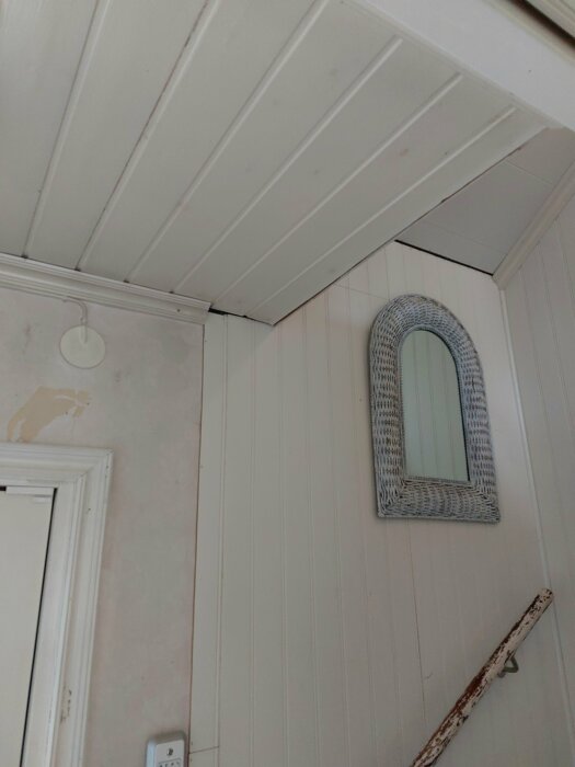 Vägg med spegel, sliten tapet; vitmålat paneltak; enkelt rum, behov av renovering.