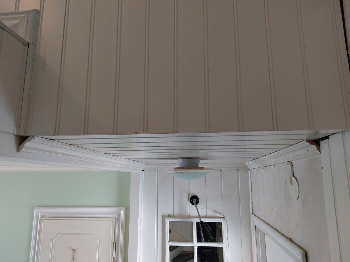 Inomhus hörnbild som visar en vit dörr, förtappning, och en enkel taklampa mot en ljus vägg.