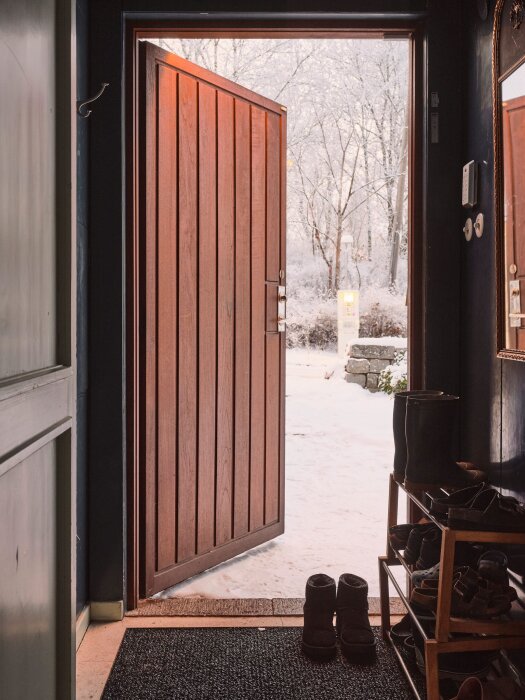 Öppen dörr från hus, utsikt över snötäckt trädgård, skohylla, inbjudande varm känsla mot kallt väder.