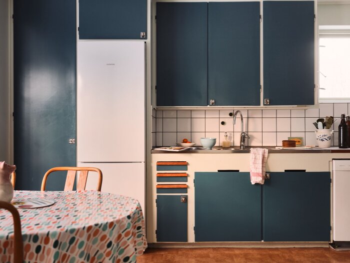 Ett mysigt kök med blå skåp, vitt kylskåp, träbord och retro inredningsdetaljer.