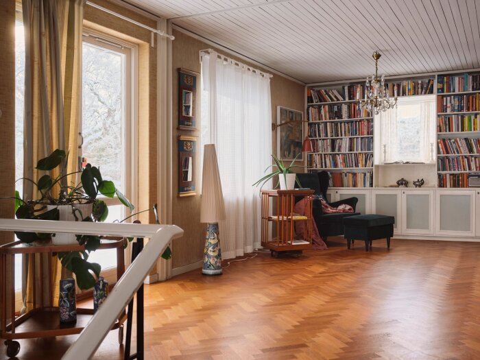 Vardagsrum med fönster, bokhyllor, trägolv, växter och klassisk inredning.