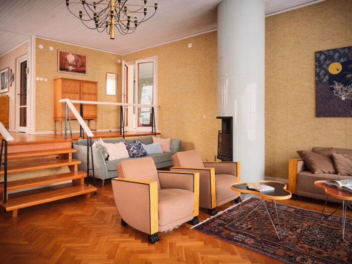 Ett vardagsrum med trägolv, kakelugn, soffor, tavlor och en trappa upp.