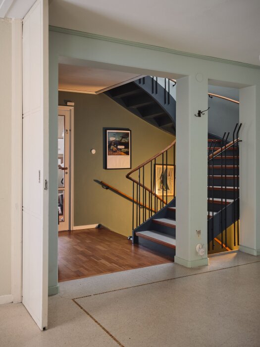Inomhusvy av ett hus, visar en trappa, konst på vägg, och trägolv genom en öppen dörrram.