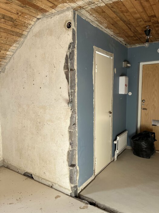 Orenoverat rum, naken betongvägg, synliga rör, trägolv, vit dörr, svart sopsäck, radiator, oavslutad renovering.