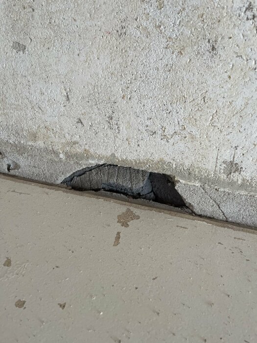 Skadat betonghörn med spricka och brutet område vid golvovergången. Slitna ytor och små fläckar synliga.