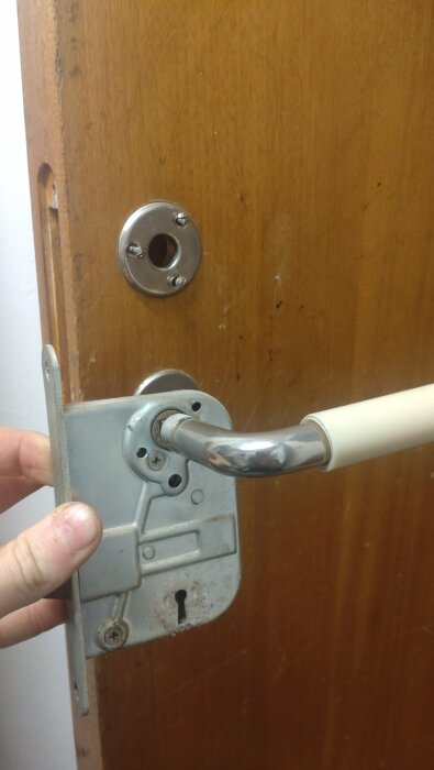Hand håller defekt dörrhandtag mot träfärgad dörr med osynligt dörrlås och rostig låsskylt.
