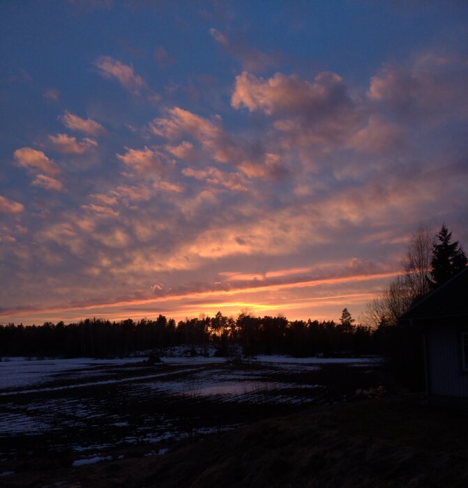 Solnedgång, molnigt himmel, orange toner, snöfläckar, träd, huskant, aftonljus, vinter till vår övergång.