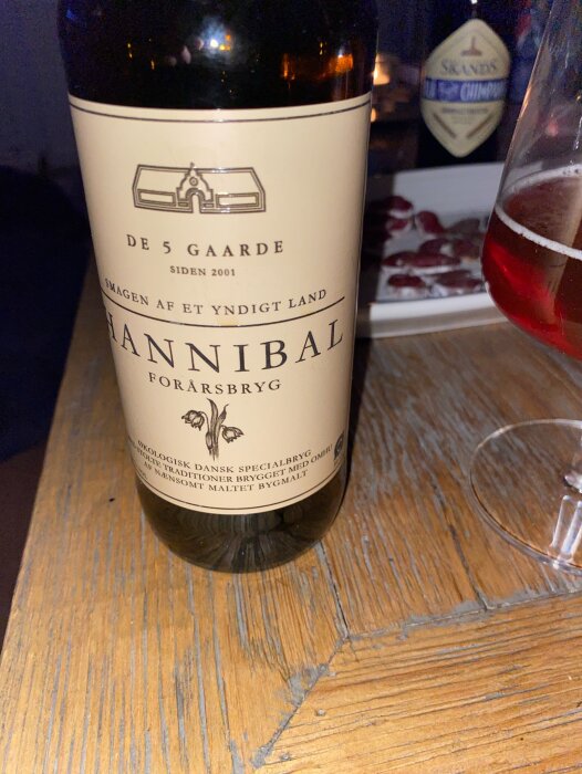 Flaska med etikett "Hannibal", skål med röd öl, skivad korv i bakgrunden på träyta.
