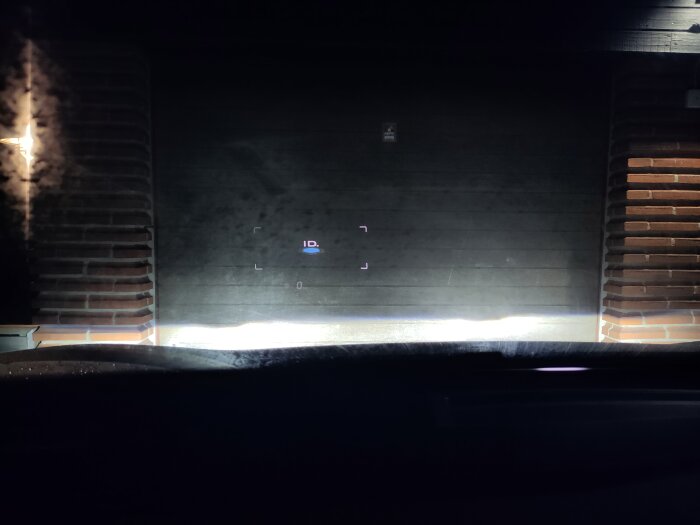 Fordonsvy inifrån, framför mörk murad byggnad, natt, belysning, head-up-display reflekteras på vindruta.