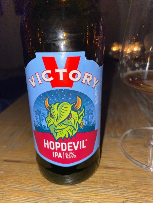 Ölflaska märkt "Victory HopDevil IPA", alkoholhalt 6.7%, på träbord med suddigt glas.