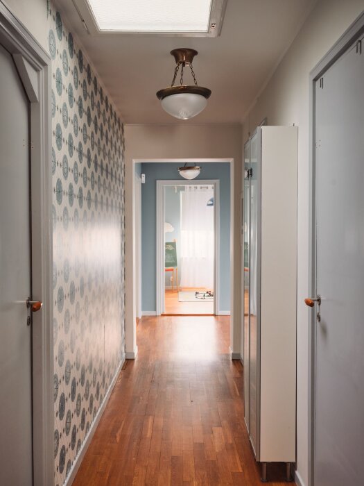 Inredningsbild, korridor med trägolv, ljusblå dörrar, vit skåp, vägglampor och mönstrad tapet.