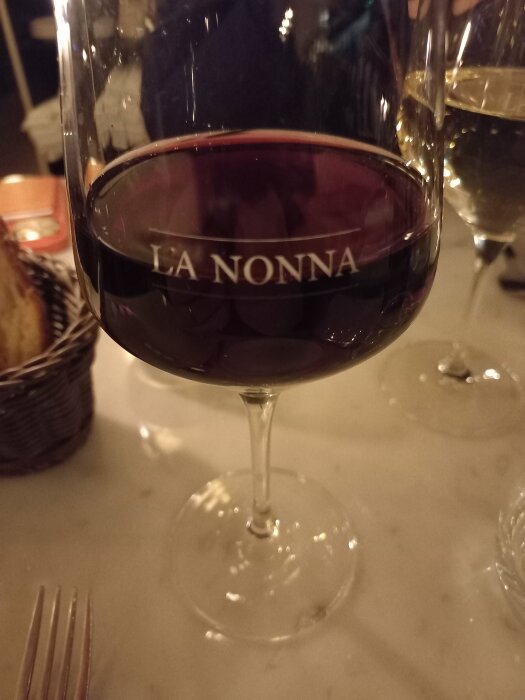 Rödvin i glas med texten "LA NONNA", reflektion, restaurangmiljö, brödkorg, andra glas, bordsduk.