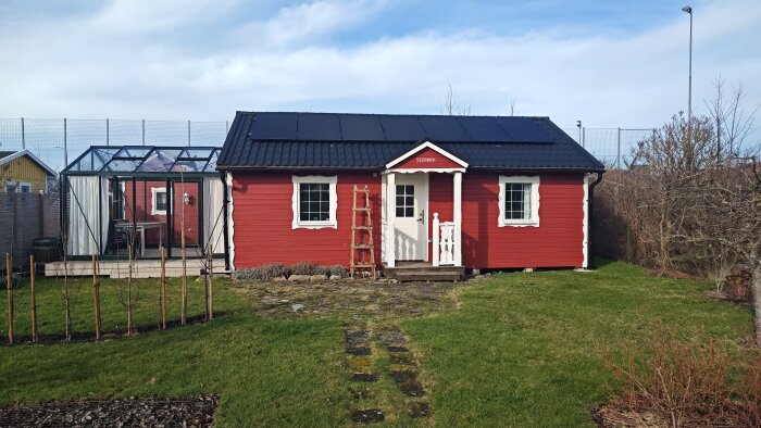 Rött hus med vita knutar, solpaneler på taket, växthus, stenlagd gång, stege, gräsmatta och klar himmel.