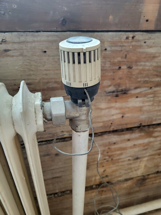 Termostatventil på radiator, trävegg i bakgrunnen, wire henger løst.