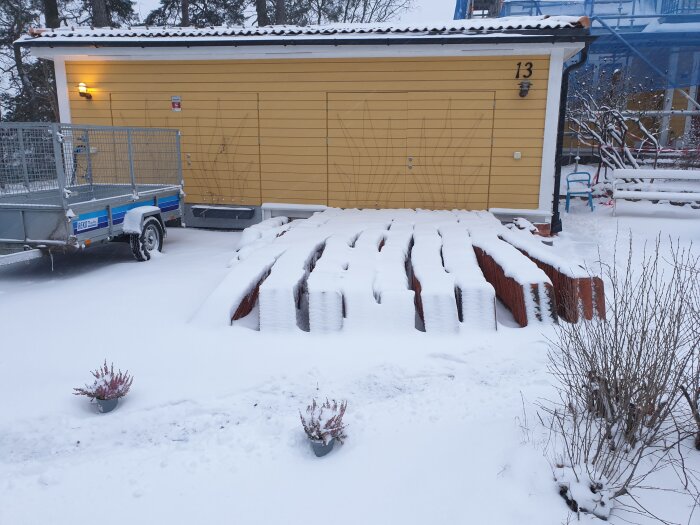 Ett snötäckt landskap med garage, släpvagn och vedstapel. Husnummer 13 synligt. Vinterstämning i ett bostadsområde.