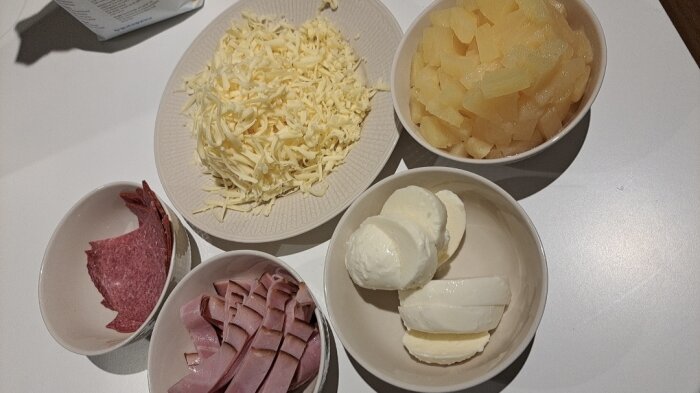 Fem skålar med ingredienser: riven ost, tärnad potatis, skinka, salami, mozzarella. Matförberedelser för matlagning.