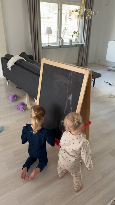 Två barn ritar på svart tavla i ett ljust vardagsrum med leksaker runt omkring.