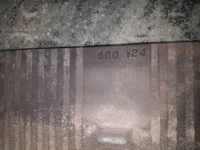 Bild på slitna metalldelar med nummer 600 124 präglat och en smutsig yta.