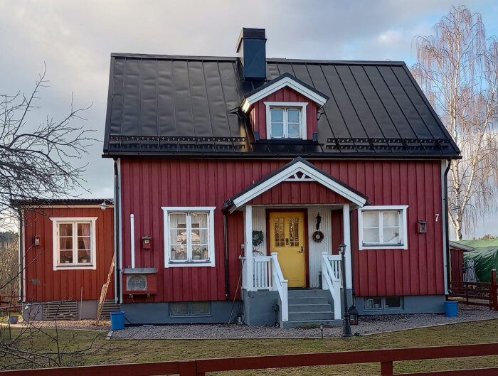 Rött hus med vita knutar, svart tak, gult dörr, trädgård, delvis molnig himmel, ingen snö, dagtid.