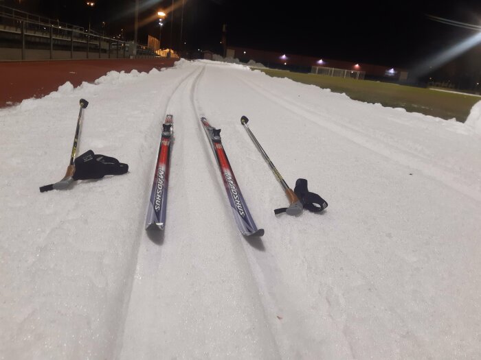 Längdskidor och stavar ligger på upplyst skidspår vid kvällstid. Vintersport, snö, utomhusaktivitet.