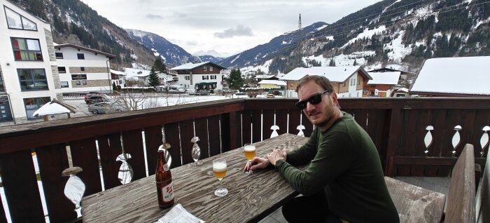 Man vid bord med dryck, blickar mot bergslandskap, snötäckta hus, moln, terrass med trästaket, avslappnad atmosfär.