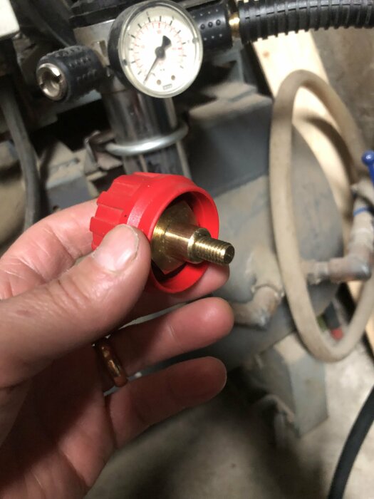 Hand håller röd mässingsventil framför kompressor med tryckmätare. Arbetsverktyg, underhåll, mekaniskt tema.