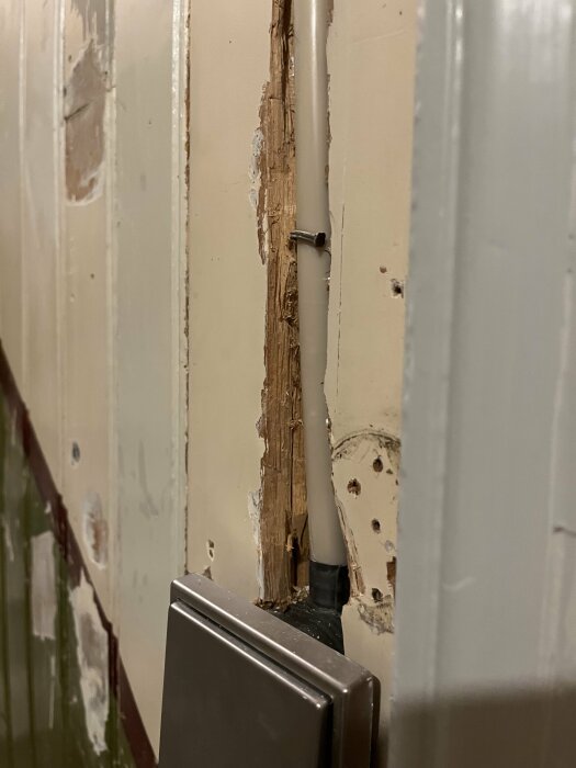 Skadad dörrkarm med flagande färg och synligt trä, nära en grå dörrstängare och vit vägg.