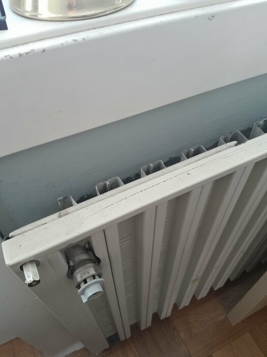 Vit radiator under fönster, termostatventil, smutsfläckar, grå vägg, trägolv, del av fönsterkarm.