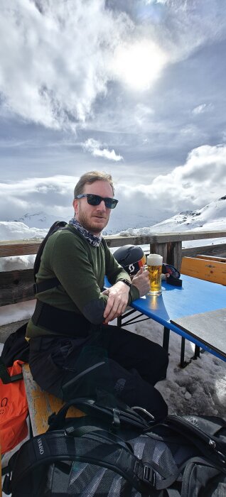 Man sitter vid bergscafé, drickandes öl, soligt, snöiga berg, ryggsäckar, vintersemester, avslappnat.