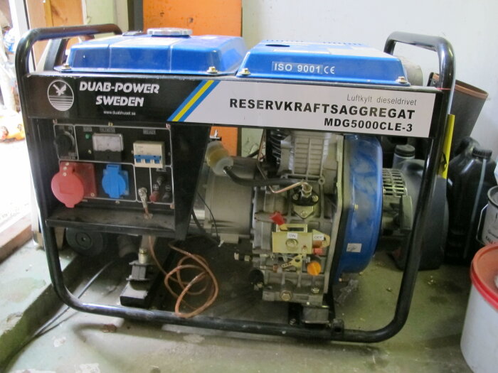 Blå och svart reservkraftsaggregat i ett garage, kabel på golvet, oljekannor bakom, text "DUAB-POWER SWEDEN".