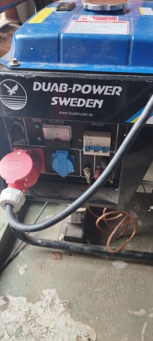 Blå generator, märke DUAB-Power Sweden, eluttag, voltmeter, säkringar, kablar, betonggolv, använd för energiförsörjning.