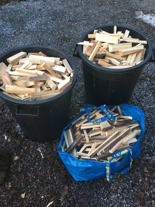 Två svarta sopkärl och en blå IKEA-väska fyllda med ved mot grusig bakgrund.