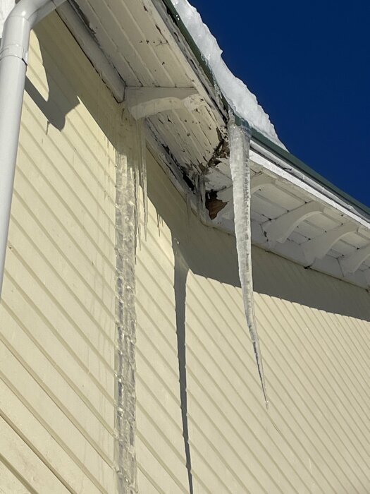 Istappar hänger från ett hus takränna under vintern; skuggor och smältande snö syns.