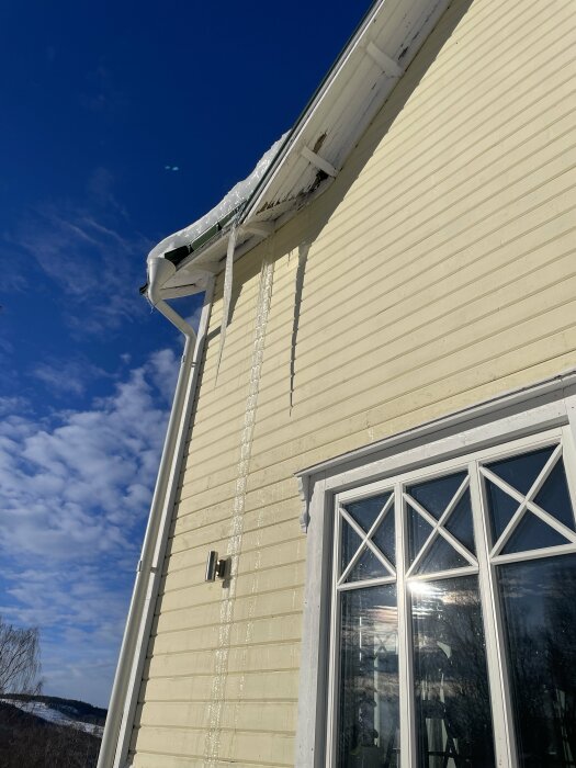 Gult hus med is istappar mot en klarblå himmel. Snö smälter, soligt väder.