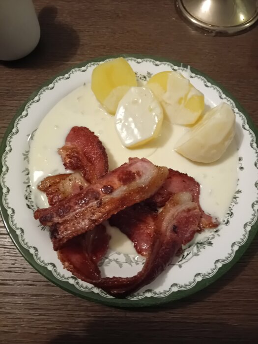 Tallrik med stekt bacon, kokta potatisar och vit sås, på ett mörkt träbord.