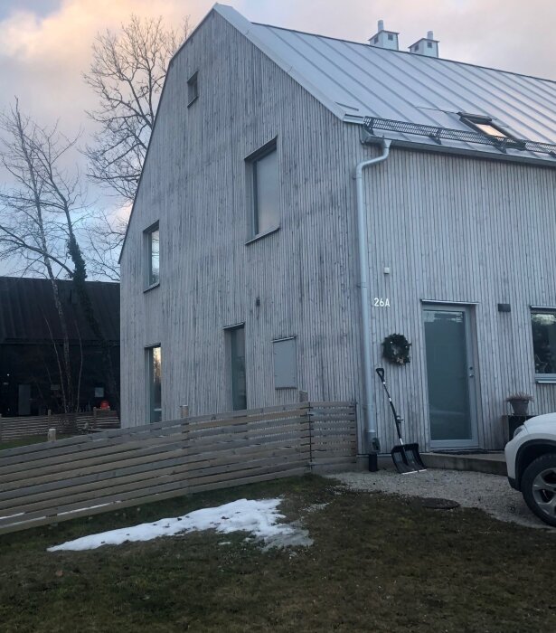 Modernt hus, träbeklädnad, stående panel, snöskyfflar, lätt snötäcke, skymning, julkrans på dörren.